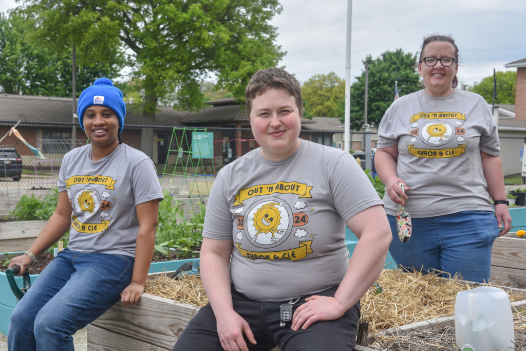 Three people in matching gray Out N' About t-shirts sit smiling at the camera while sitting in the community garden with green trees and a cloudy sky behind them