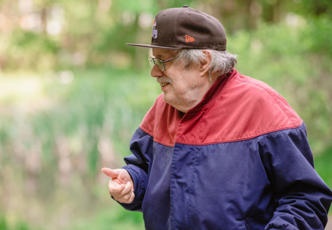 Close up of a profile view of a senior in a jacket wearing a brown baseball hat with blurred greenery in the background