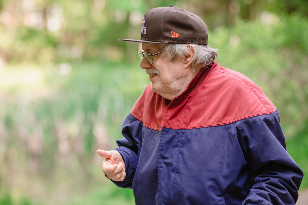 Close up of a profile view of a senior in a jacket wearing a brown baseball hat with blurred greenery in the background