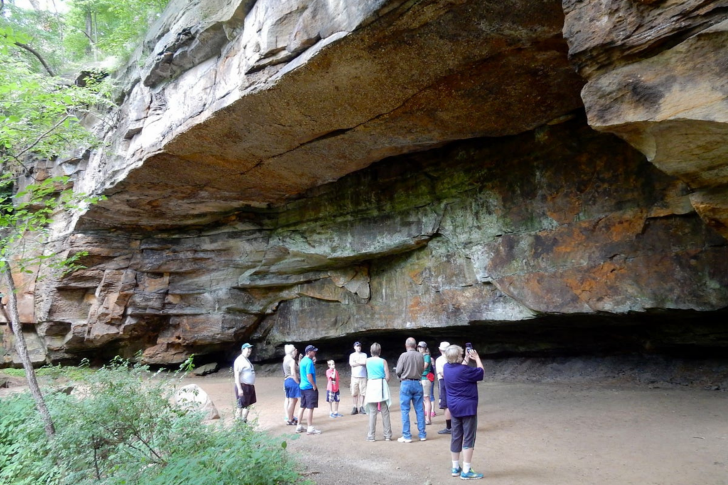 visitors on a guided hike stand below a prominent rock shelter in Gorge Metro Park