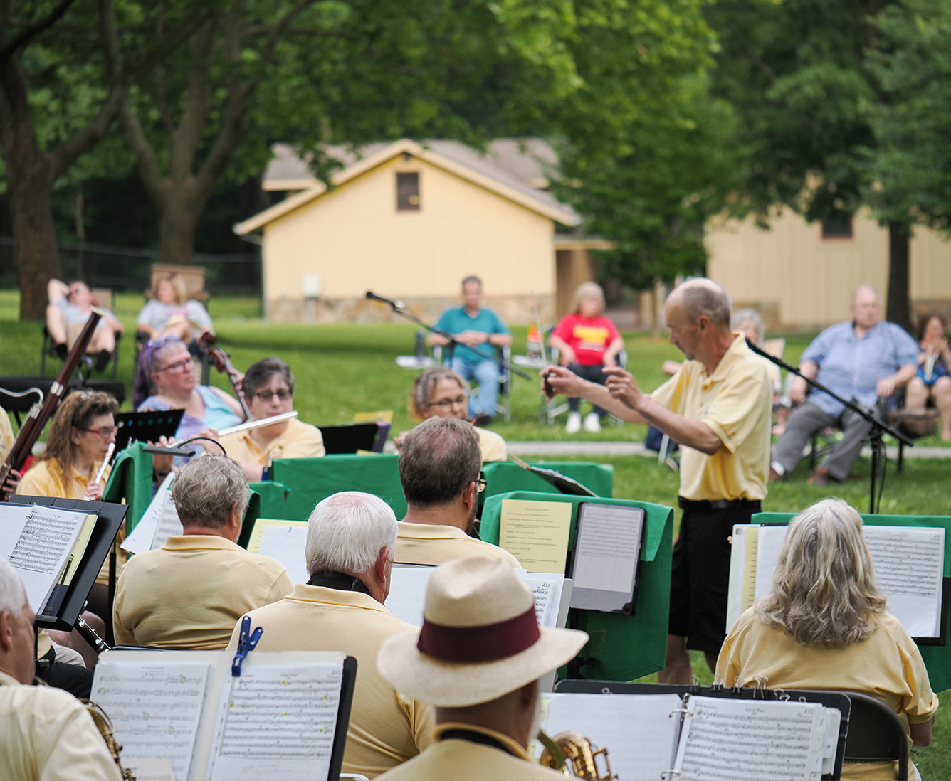A man conducts a band outside as people sitting in lawn chairs watch.