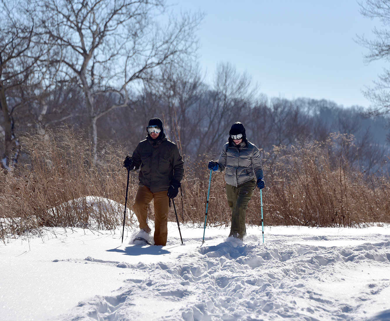 Two people bundled up in winter coats, hats and goggles snowshoeing on an open trail.